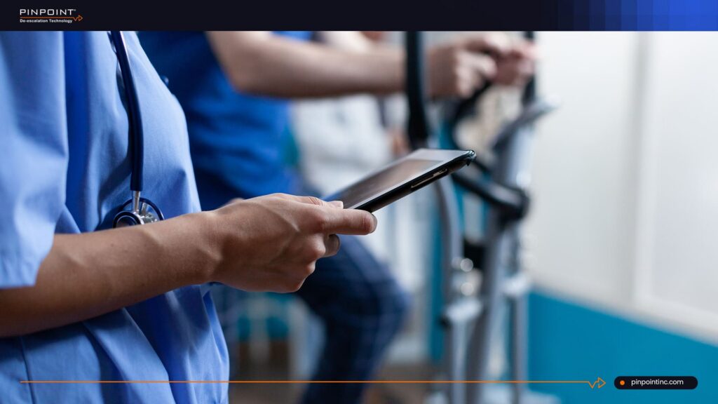 A close-up view of a healthcare professional in blue scrubs holding a digital tablet in a medical facility, with hospital equipment blurred in the background.