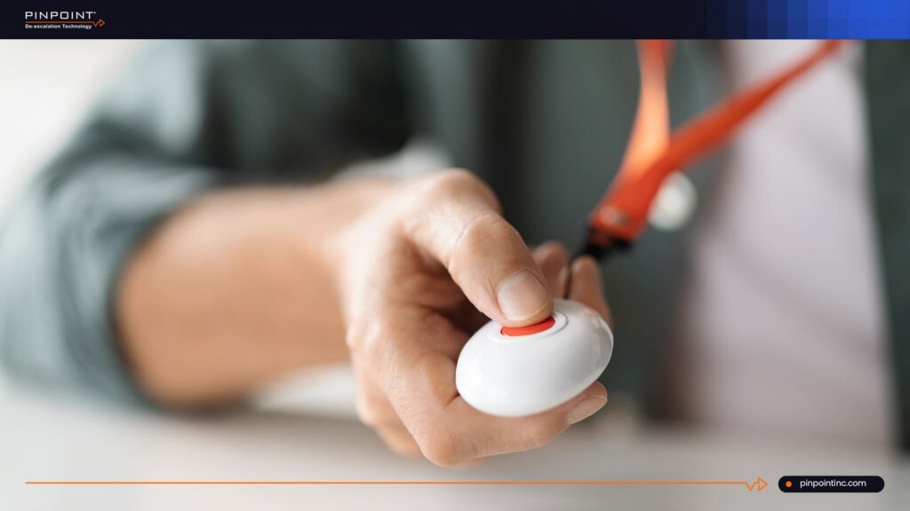 A close-up shot of a staff member's hand pressing a red button on a white, ergonomic wearable panic device attached to an orange lanyard.
