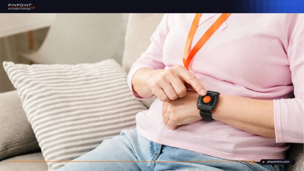 A close-up of a healthcare worker sitting on a couch and pressing a prominent orange "CALL" emergency duress button worn on a black wristband strap.