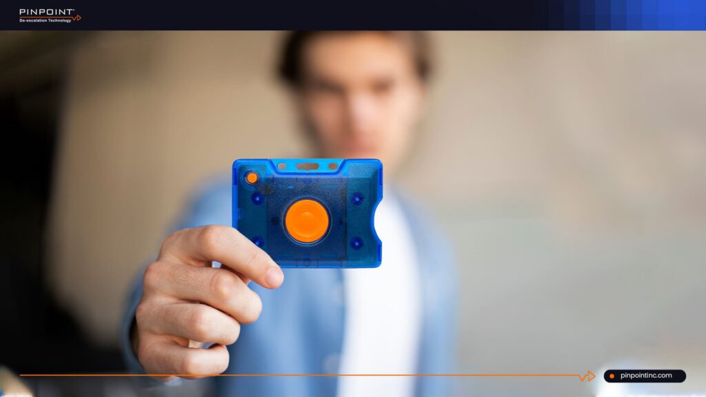 A close-up of a person's hand holding a blue semi-transparent Pinpoint duress panic badge with a large orange button in the center. The background features a blurred young man looking forward.