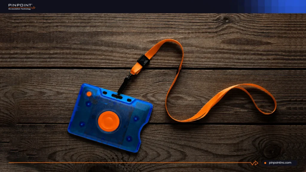 A blue semi-transparent Pinpoint duress safety badge with a central orange button, connected to an orange lanyard strap, lying flat on a dark textured wooden plank surface.