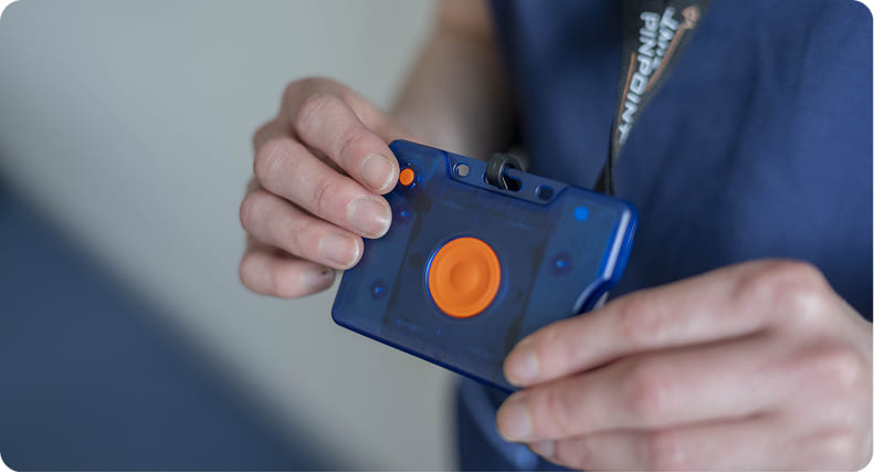 A close-up view of hands holding a blue translucent wearable staff safety duress badge featuring a large orange wireless panic button.