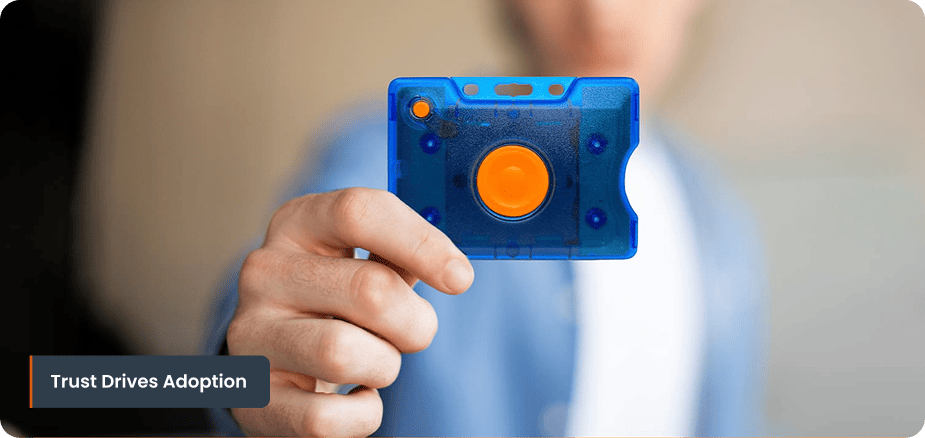A female registered nurse wearing blue scrubs presses a wearable wireless panic button badge pinned to her pocket inside a hospital corridor, triggering an active alert system visible on a background wall monitor.